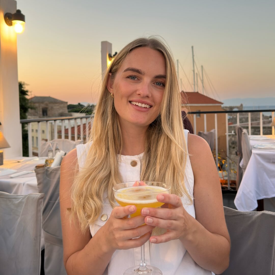 Marisha sidder ved et restaurantbord på en udendørs terrasse ved solnedgang med en drink